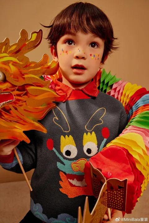 A child wears a dark dragon-motif jumper with a red collar, beside a colourful paper dragon.